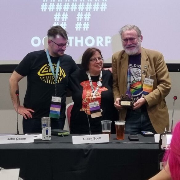 John, Alison, and Nicholas Whyte stand in front of a projection of the Octothorpe podcast and behind a panel table. Each of them wears a convention badge, and Nicholas holds the Glasgow Landing Zone Rocket. Nicholas is looking at the camera, while John and Alison are not quite as good at this. The table they stand behind holds beers, coffees, convention newsletters, phone batteries, microphones, and table tents.