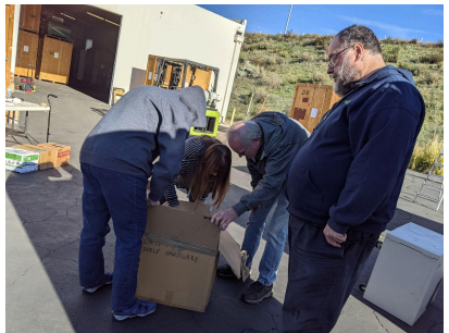 Color photo. Taken outside storage unit. Four people looking into cardboard storage box.